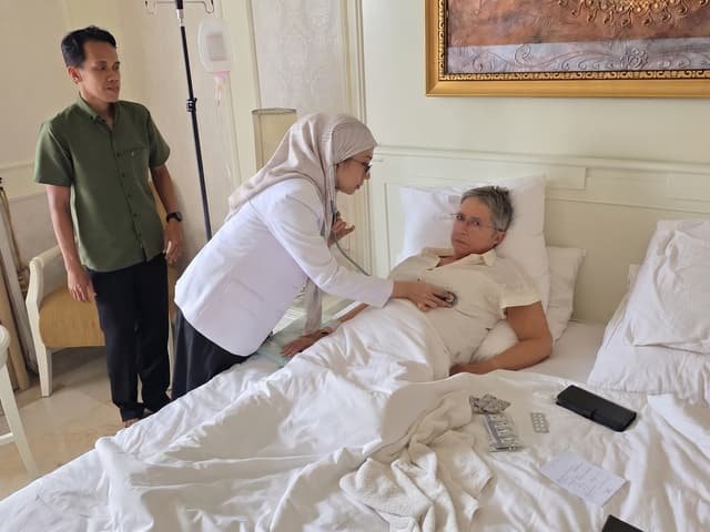 A Mitra 911 doctor examining an international guest with a stethoscope in her hotel room, an IV line set up beside the bed